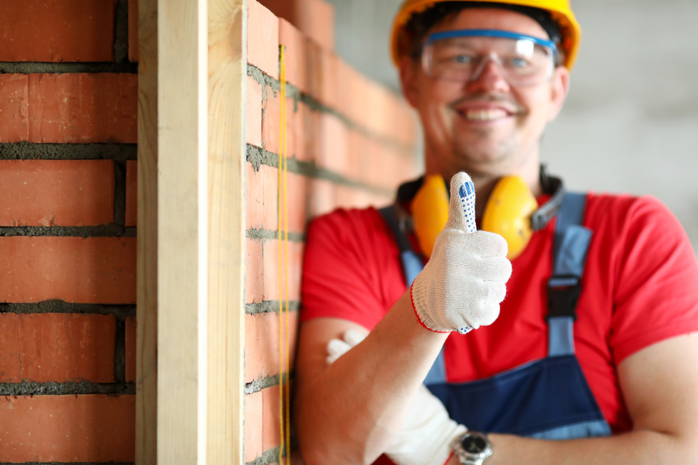 Trabajador de la construcción con casco y guantes junto a pared de ladrillos, mostrando un pulgar hacia arriba.